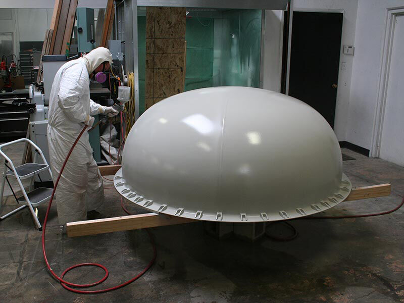 Image of manufacturing area with a large dome-shaped radome in the center, elevated on wood posts. A technician is wearing protective gear and is spray painting the large belly radome with an off-white coating.